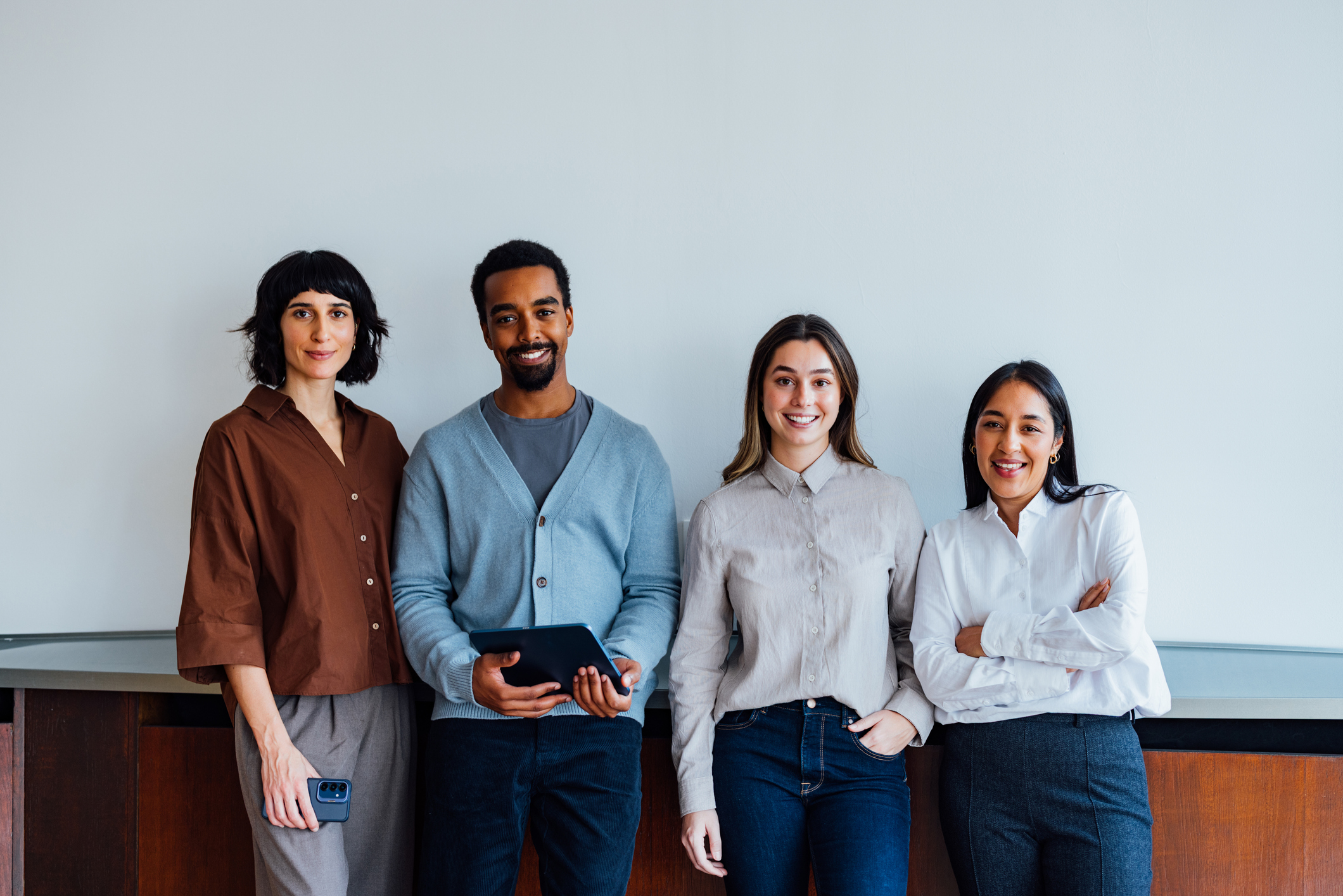Four diverse professionals stand together, smiling against a light wall, showcasing teamwork and friendly collaboration.